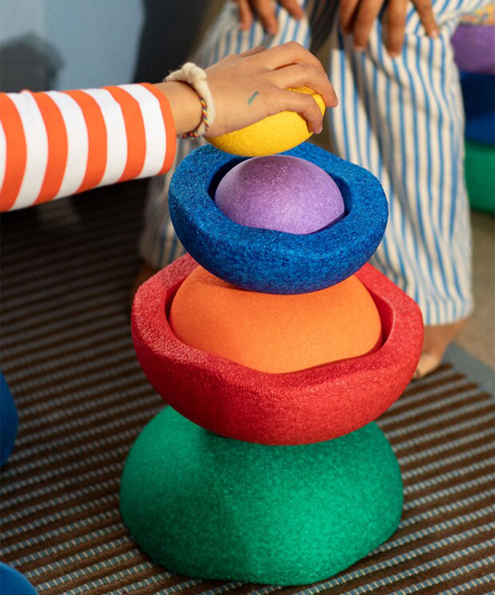 A hand stacks Le Inside Rainbow’s colorful, textured foam shapes on a striped mat, with another person nearby.