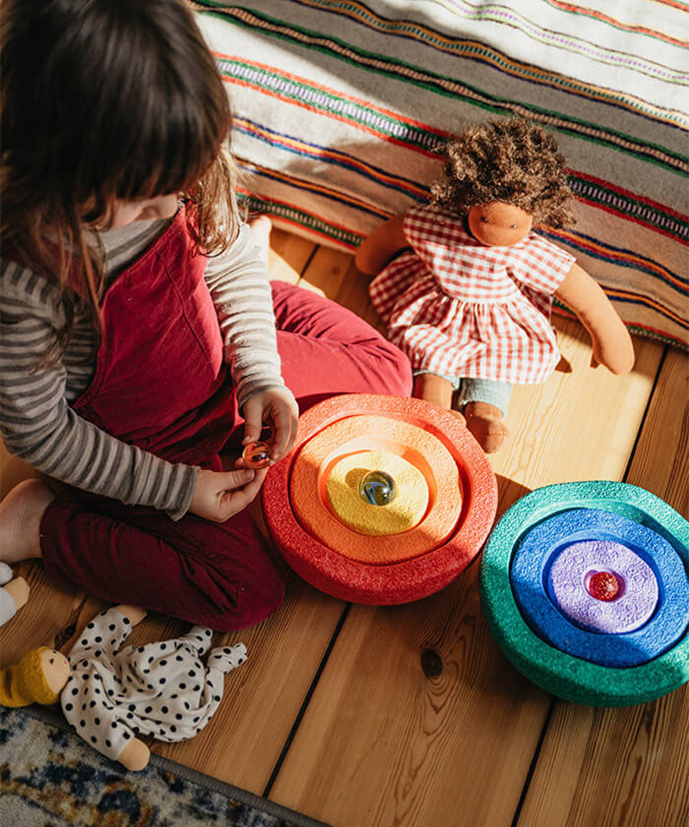 A young child plays with the Le Inside Rainbow stacking toys on a wooden floor, surrounded by two dolls and sunlight.