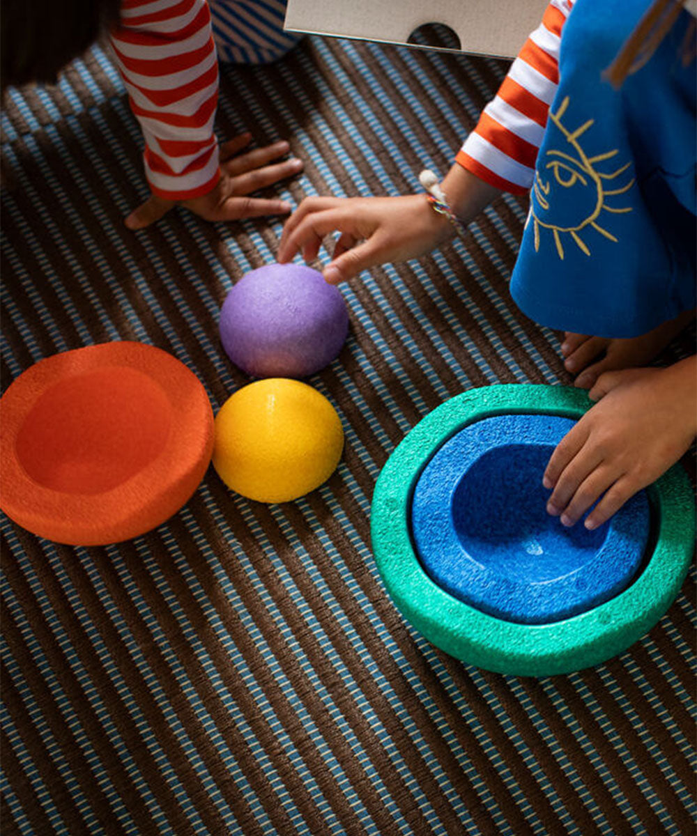 Children’s hands reach for Le Inside Rainbow’s colorful textured bowls and balls on a striped carpet.