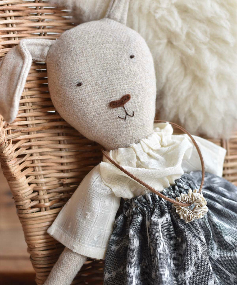Ruthie le Rabbit - Ikat Skirt, a fabric bunny doll in a white blouse, patterned skirt, and necklace, sits on a wicker chair.