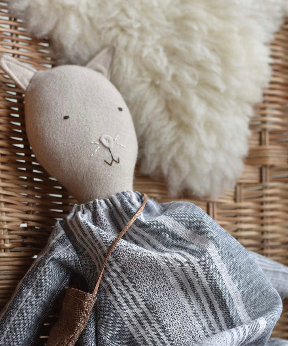 Mae le Barn Cat, wearing a striped linen dress, rests on a wicker chair topped with a fluffy white cushion.