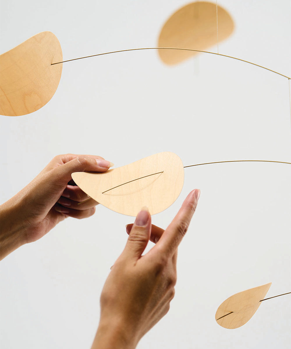 Two hands assemble the Le Drifting Clouds wooden mobile, featuring leaf-shaped pieces on thin wires, against a plain background.