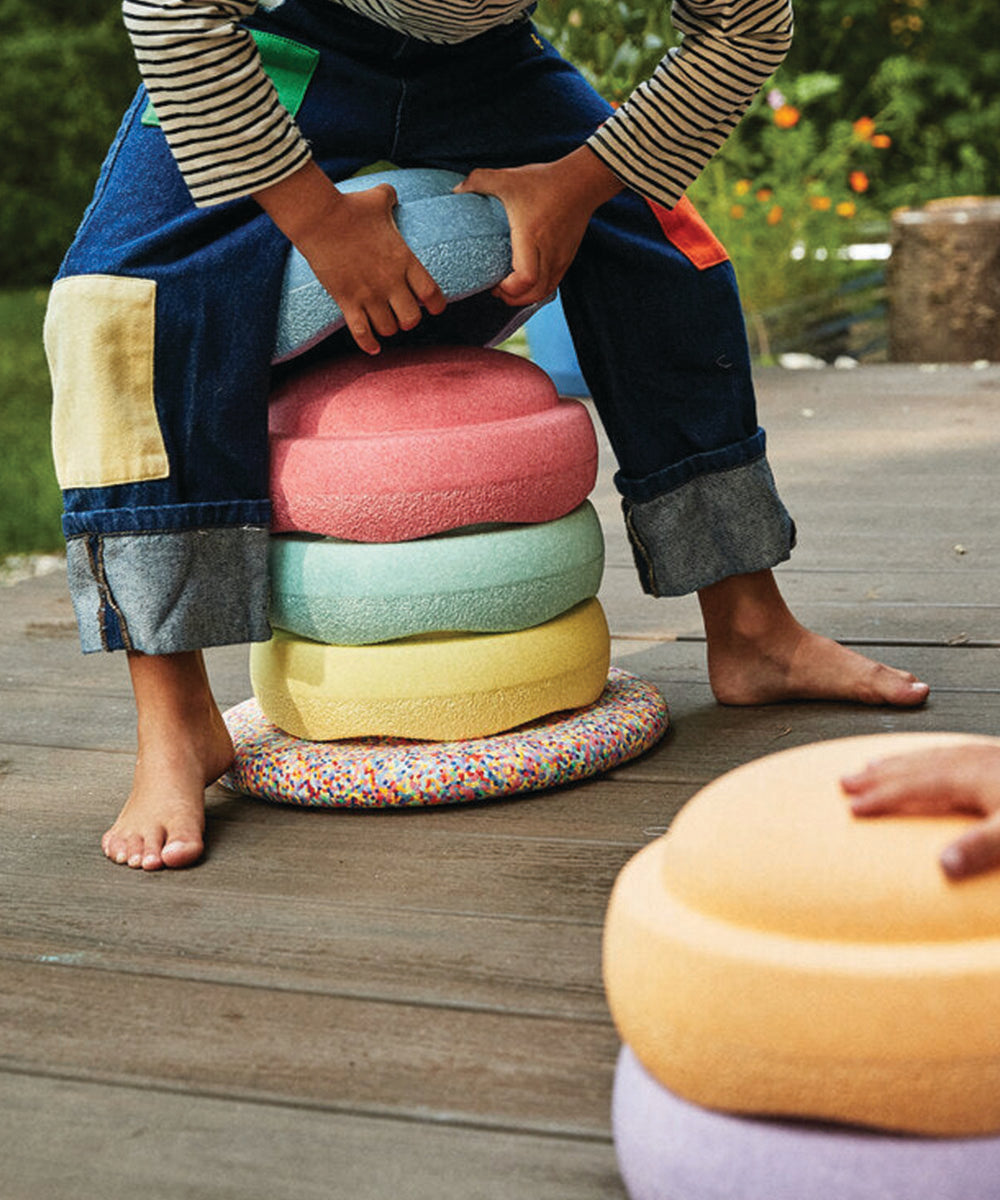 A child arranges the Rainbow Set's colorful, soft cushions on a wooden deck, barefoot and in patched jeans.