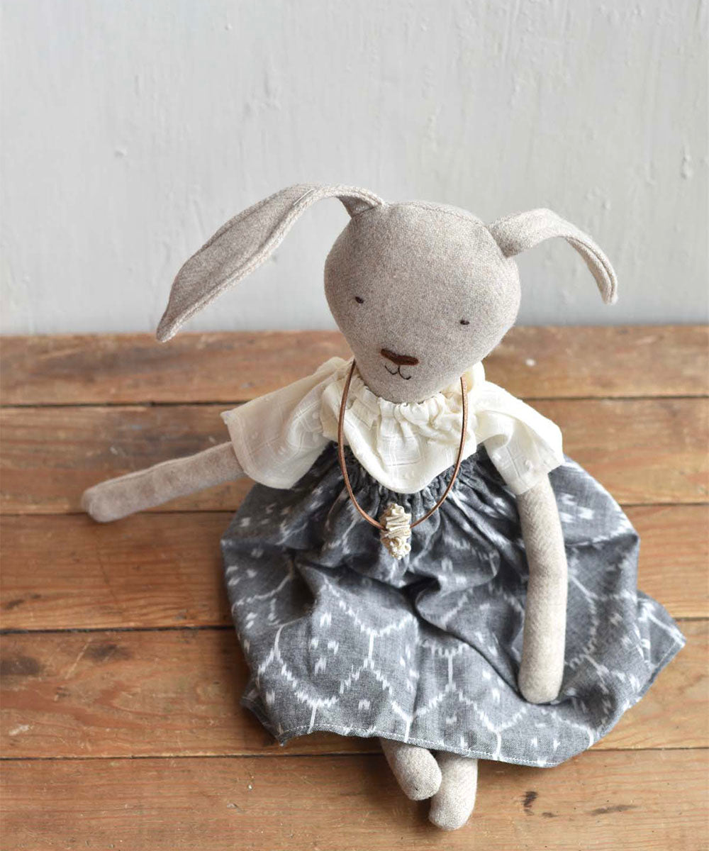 Ruthie le Rabbit - Ikat Skirt: A stuffed fabric bunny in a white shirt, gray patterned skirt, and necklace, sits on a wooden surface.
