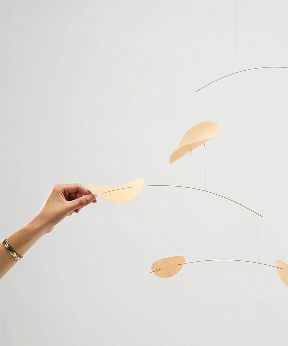 A hand touches the Le Drifting Clouds mobile, featuring curved, leaf-shaped wooden pieces suspended on thin wires.
