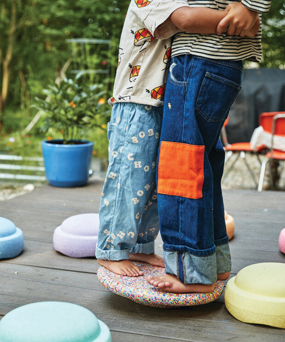 Two children stand barefoot outdoors, hugging each other closely on the Rainbow Set balance cushion.