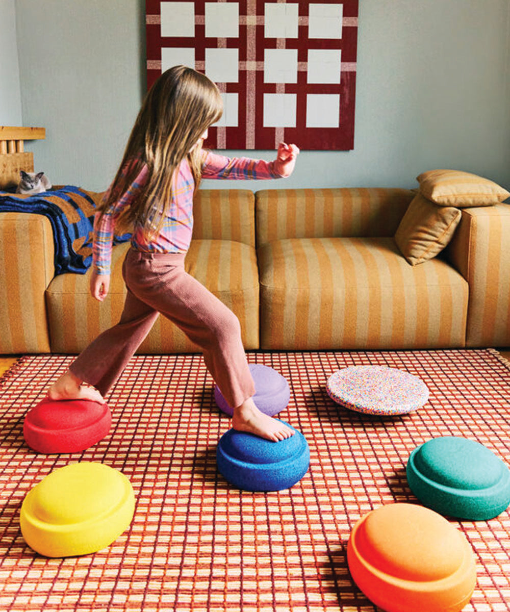 A young girl balances on the Rainbow Set stepping stones in a living room with a yellow striped sofa and patterned decor.