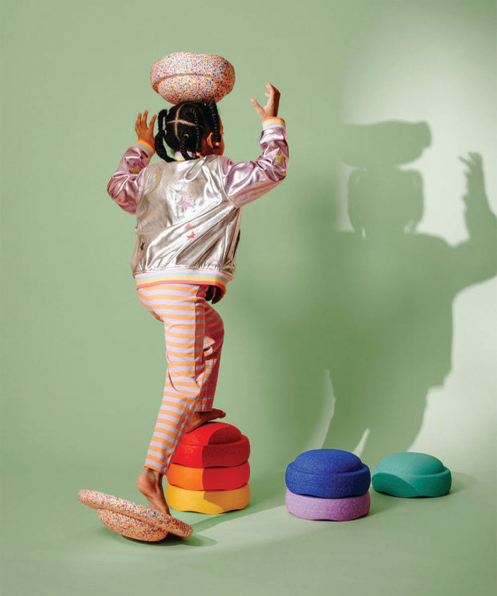A child balances the Complete Set cushions on their head and steps on stacked cushions, with a green background.