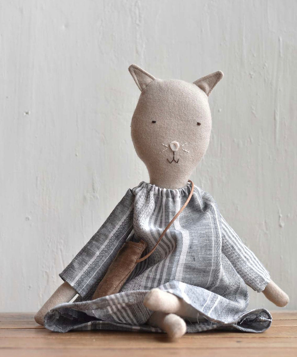 The Mae le Barn Cat, dressed in a striped linen dress, sits on a wooden surface in front of a light textured wall.