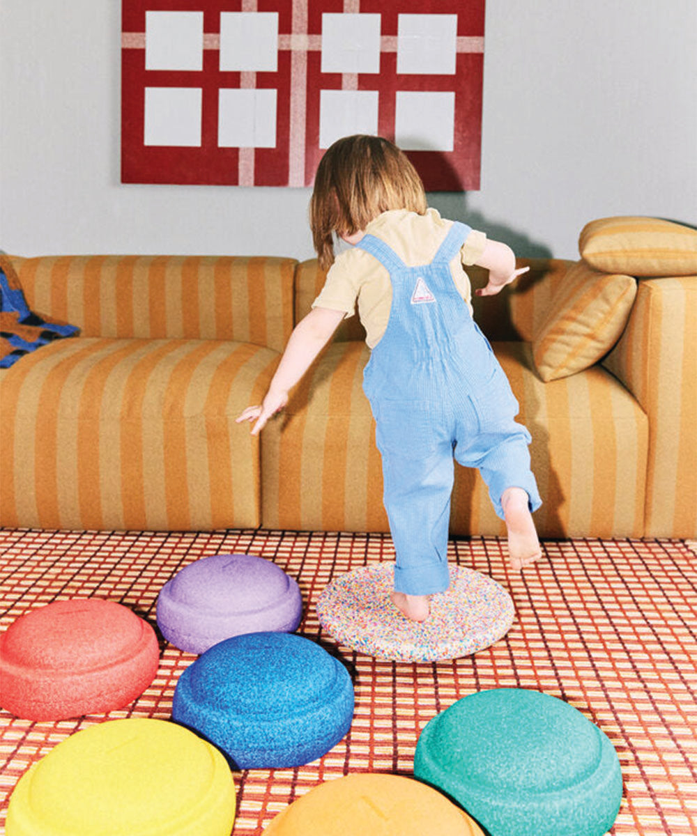 A child plays on the Rainbow Set of colorful stepping stones in a living room with a yellow striped sofa.