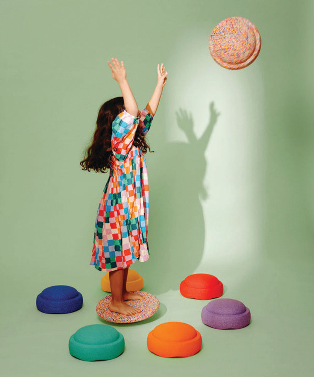 A young girl in a colorful dress throws the Complete Set cushion, surrounded by vibrant cushions on a green background.