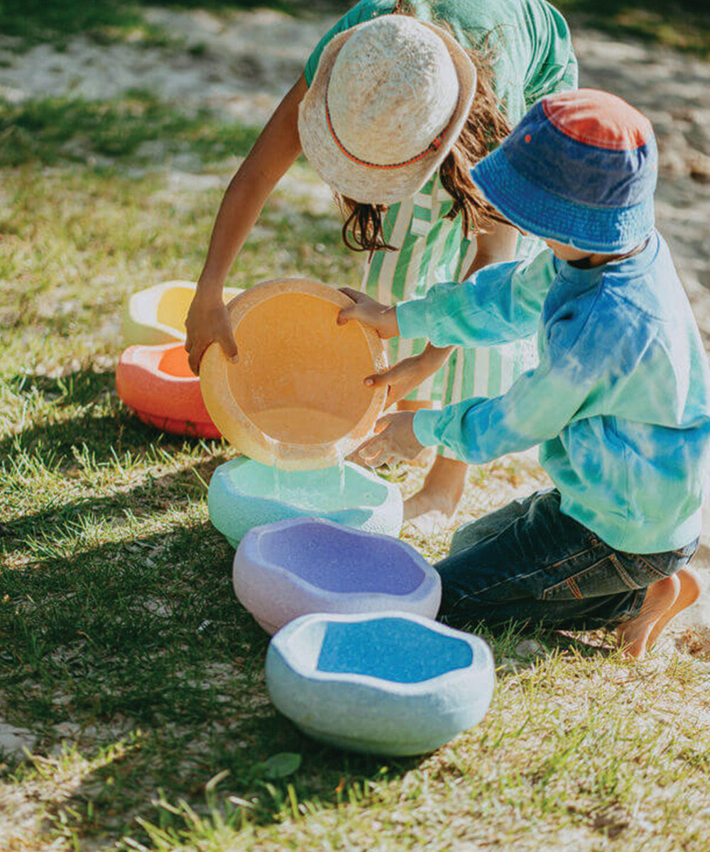Two children pour water into colorful Complete Set bowls arranged in a row on the grass outdoors.