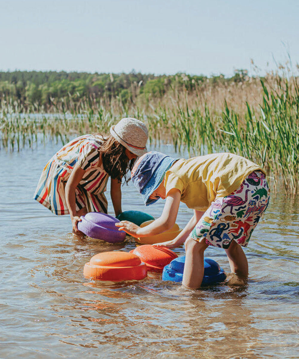 Two children in sun hats play with the Complete Set on colorful stepping stones in shallow water near tall grass on a sunny day.
