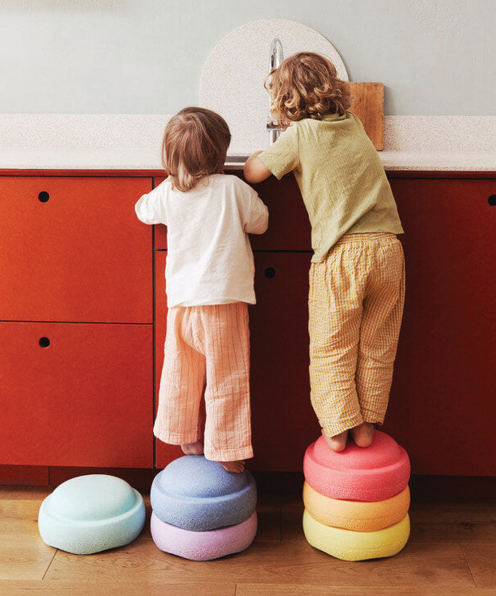 Two young children stand on the Complete Set of colorful foam stools at a red kitchen counter, reaching the sink.