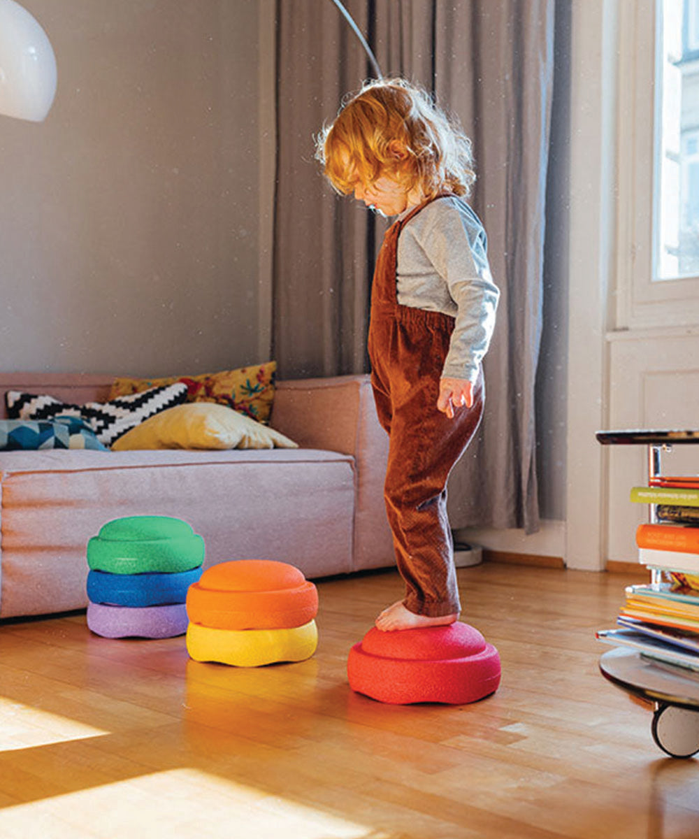 A young child balances on the Complete Set’s colorful stacked cushions in a cozy, sunlit living room.