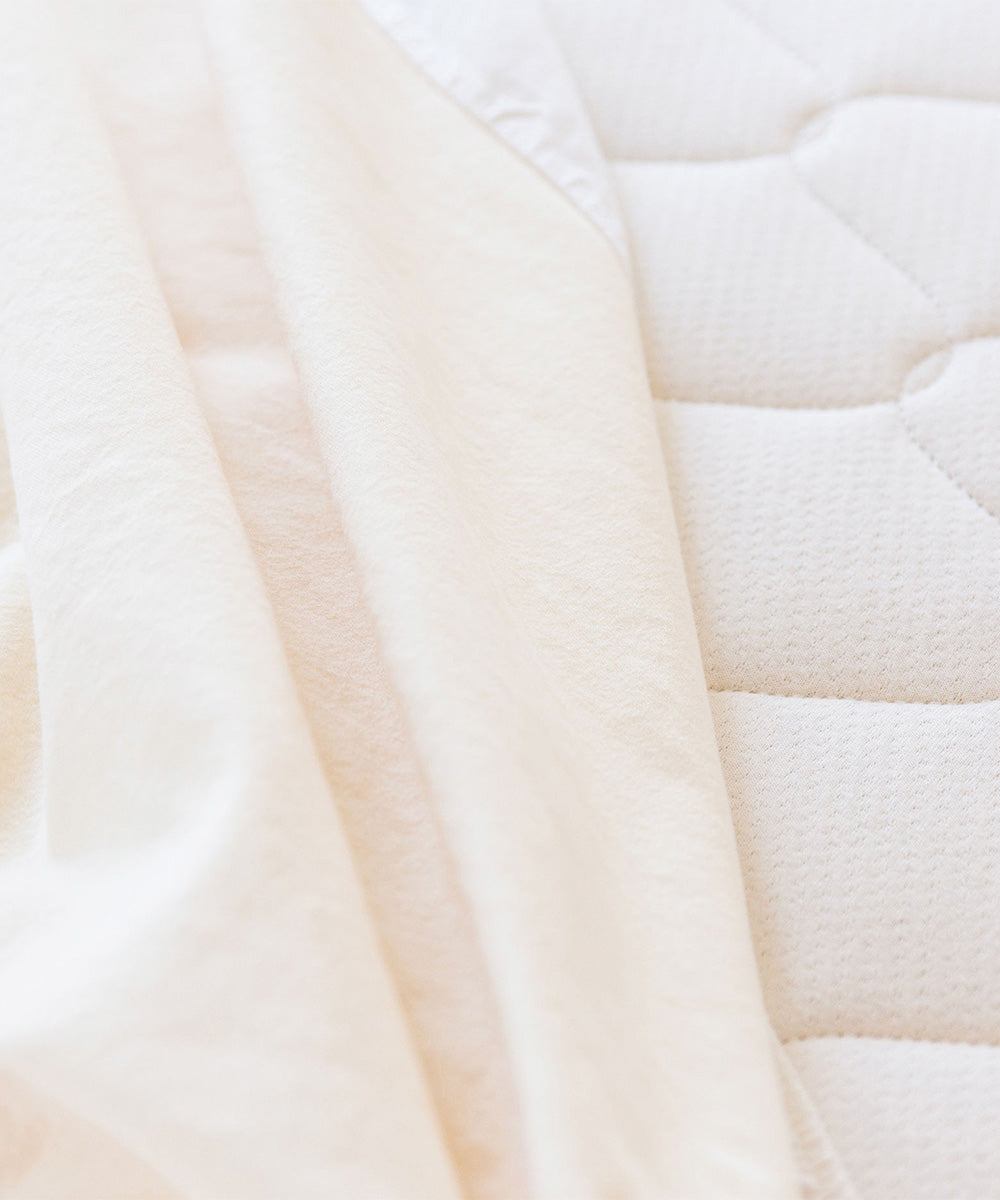 Close-up of the Organic Waterproof Kids Mattress Protector Pad in cream, partially covering a textured quilted mattress surface.