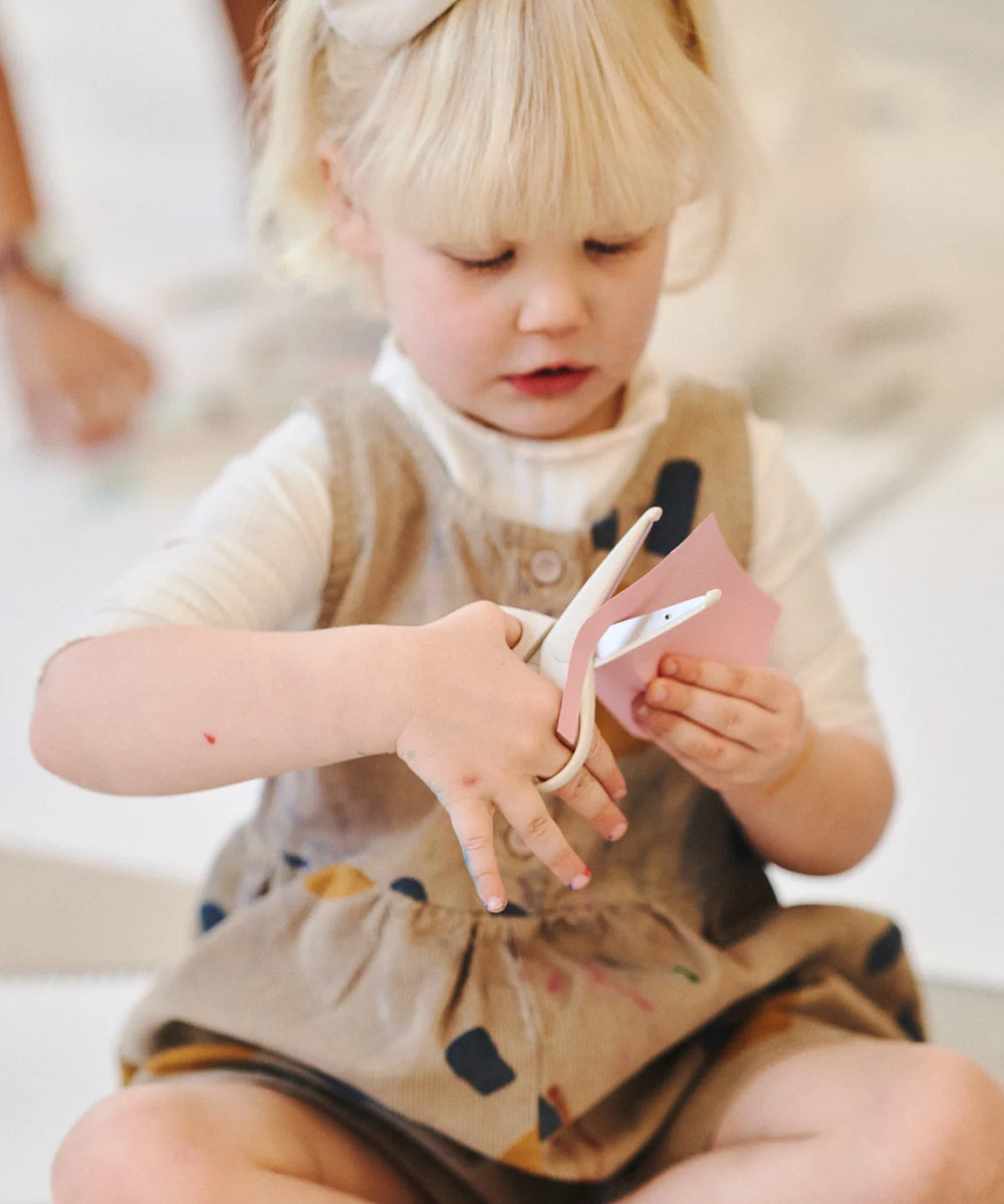 A young girl with blonde hair uses Le Gentle Scissors to cut pink paper while sitting on the floor.