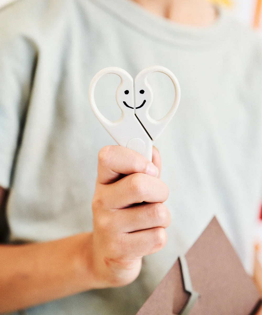 A person holds Le Gentle Scissors, featuring a smiling face design, near a piece of brown paper.