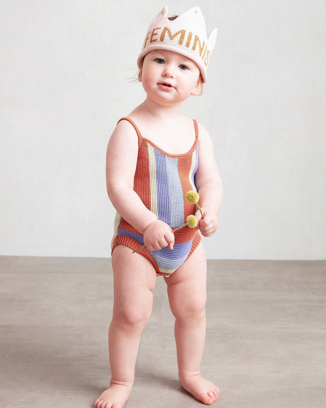 A toddler wearing a colorful striped outfit and a Feminist Crown stands on a gray floor.