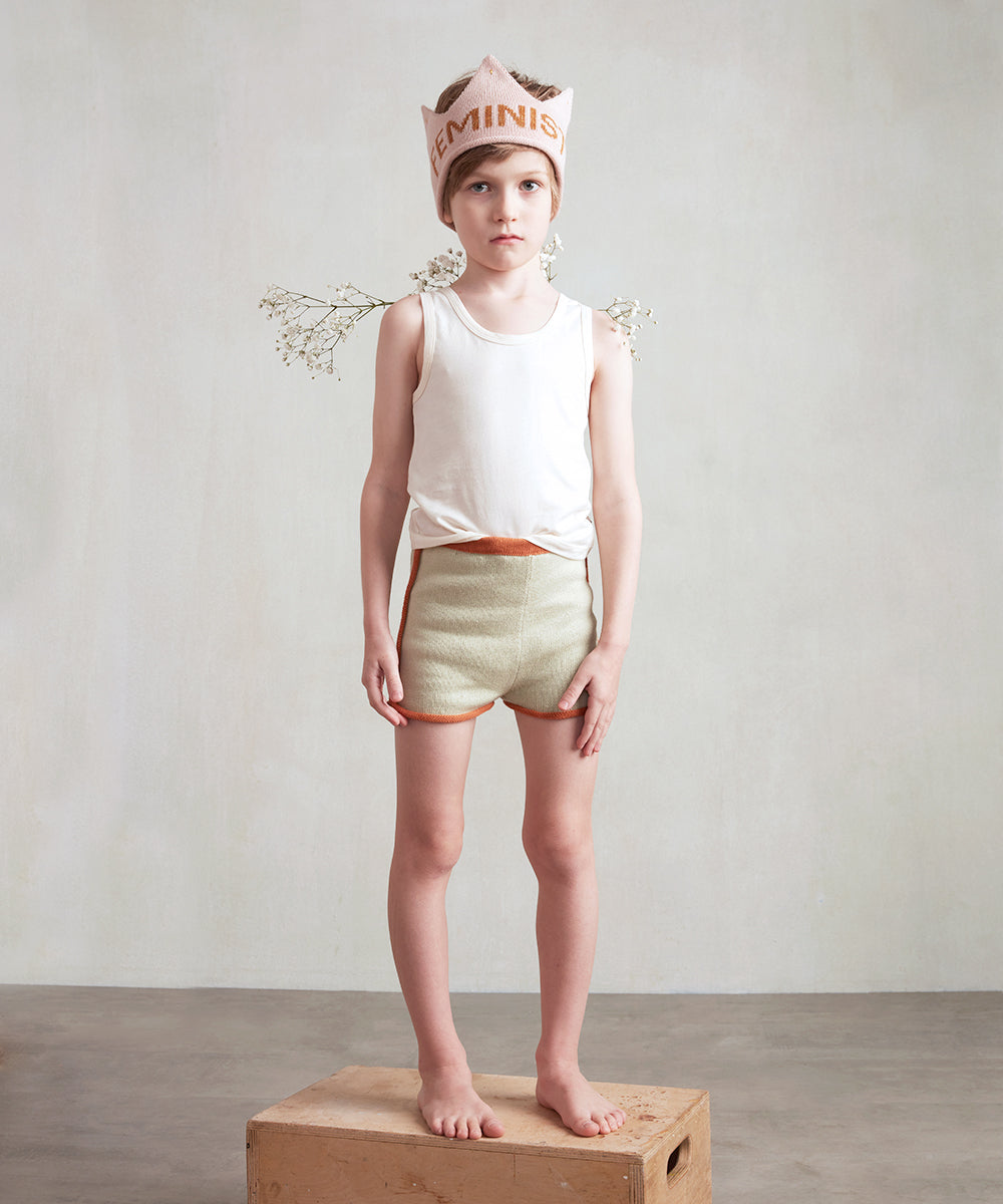 A child in a tank top and shorts stands on a box, wearing the "Feminist Crown" and decorative wings.