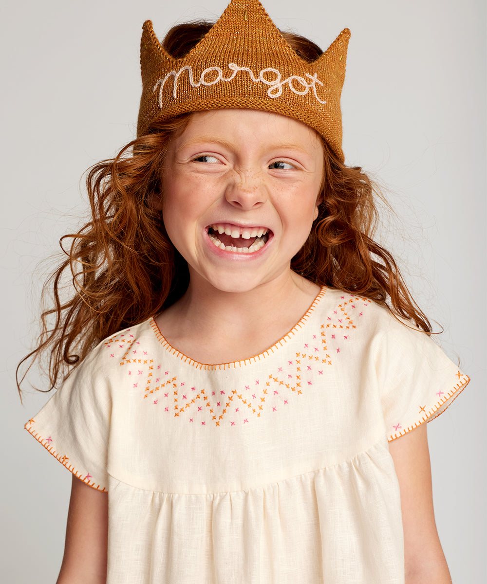 Smiling girl with red hair wears a Personalized Gold Crown in Light Pink labeled "Margot" and a cream dress with colorful stitching.