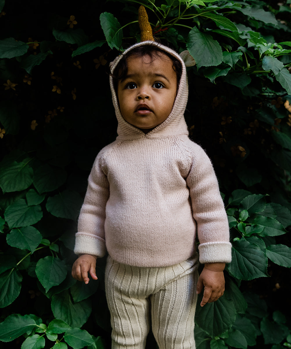 A toddler wearing the Unicorn Hoodie stands in front of lush green foliage, gazing upward.