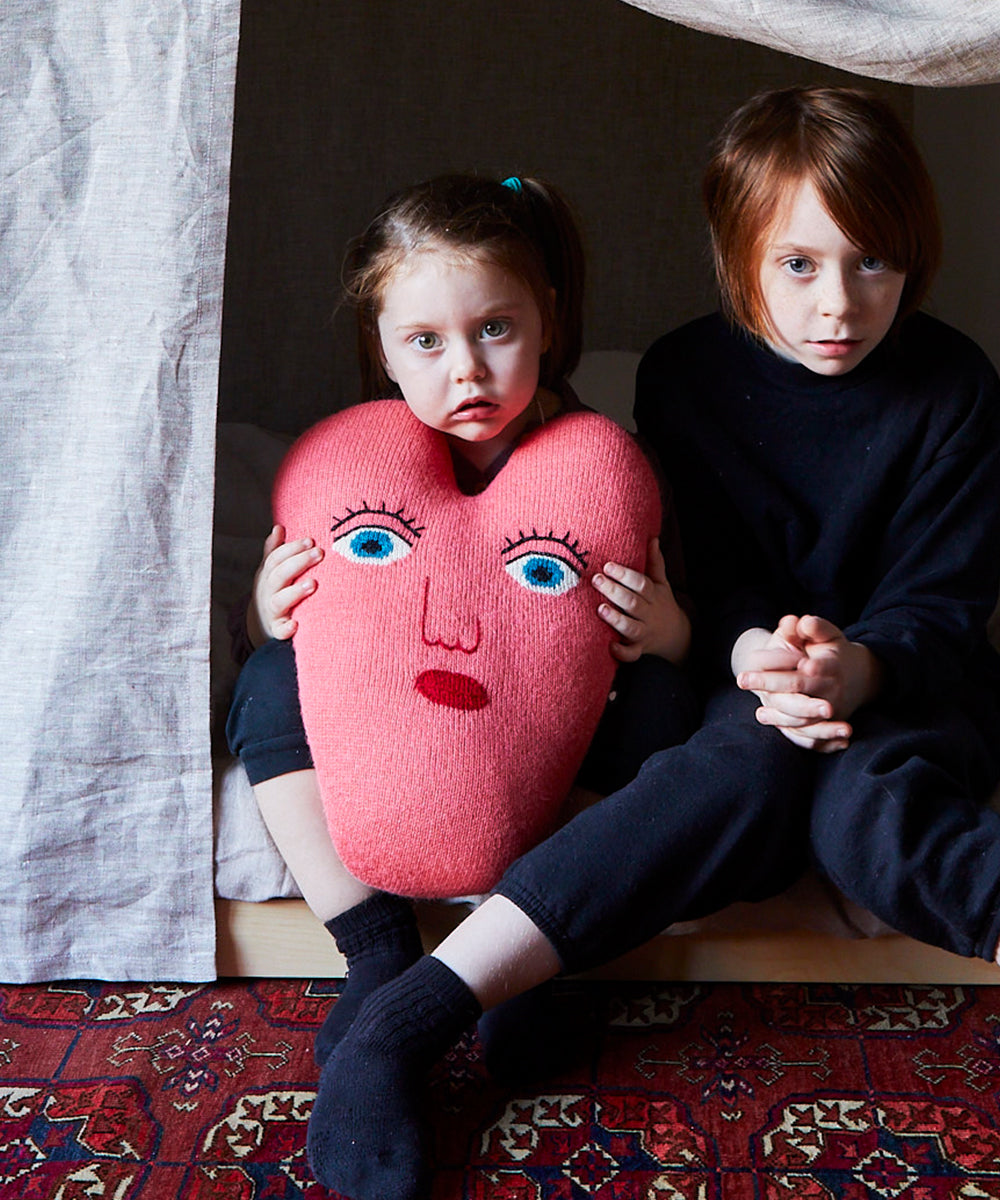 Two children sit on a rug inside a tent. One holds a pink heart-shaped pillow with a face design.