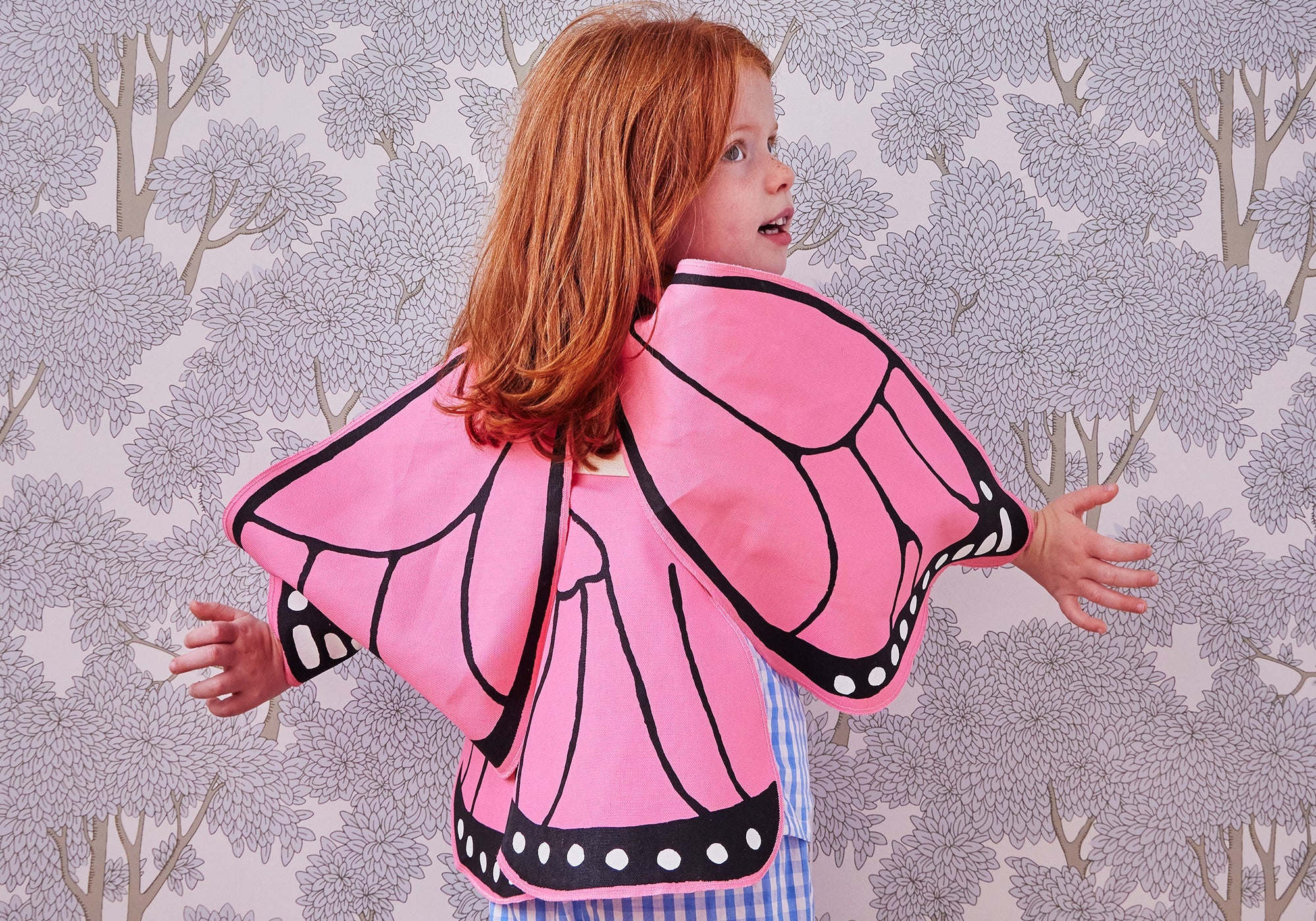 Child with red hair wearing pink butterfly wings, standing against a floral wallpaper background.