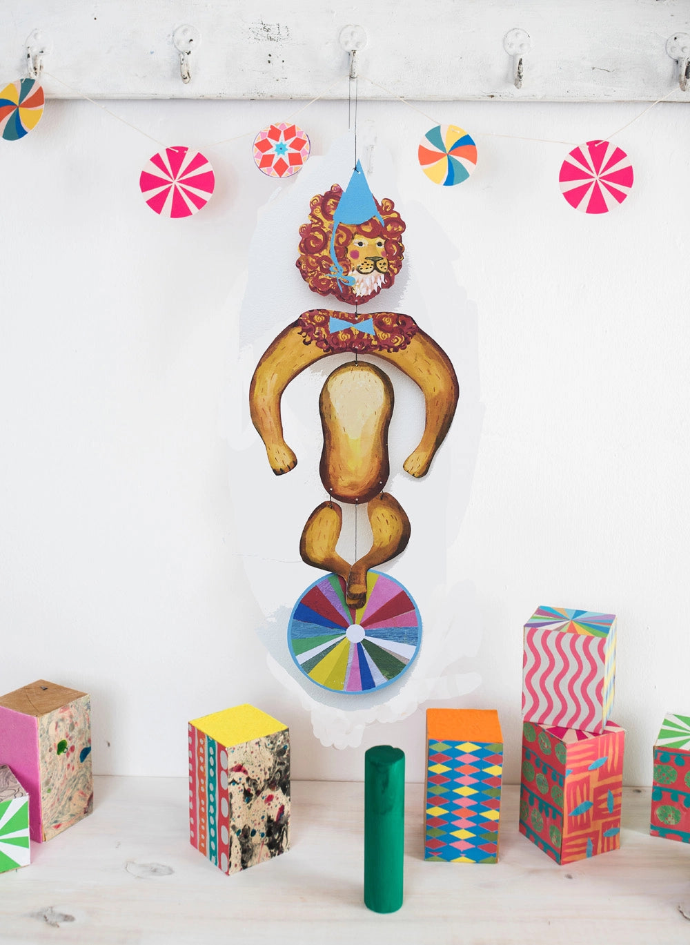 The Le Mobile - Circus Lion, a paper lion on a colorful wheel, hangs above patterned blocks and bright paper pinwheels.