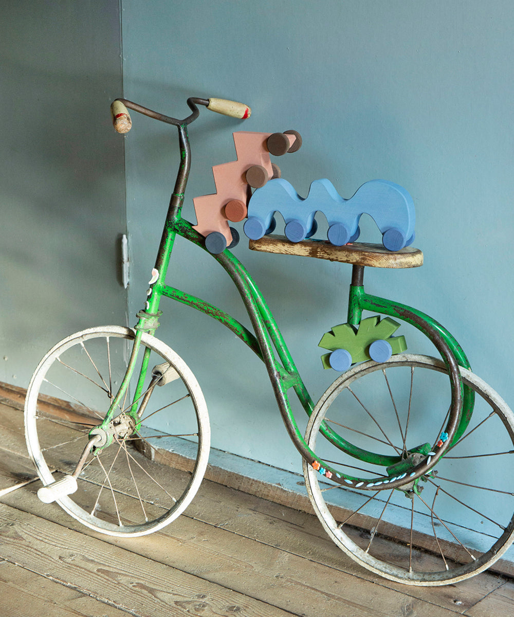 The Le Zigzag Vehicle features a vintage green tricycle adorned with wooden toy cars and dinosaurs on its seat and wheels.