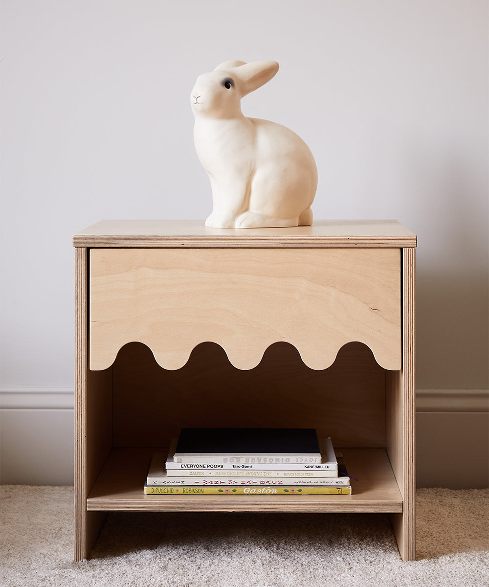 A wooden nightstand with a scalloped edge drawer holds three books on the lower shelf and features the Le Bunny Lamp on top.