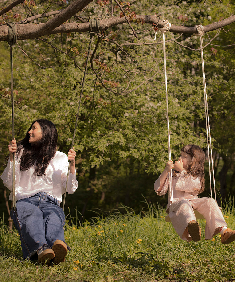 Two people sit on Le Wooden Swing hanging from a tree branch in a lush, grassy area with trees in the background.