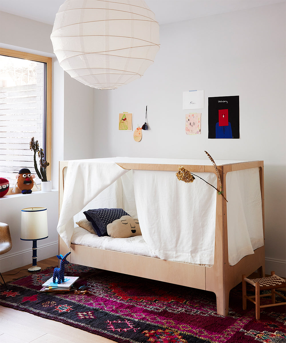 A childrens room featuring the Perch Nest Bed, a delicate white canopy, a vibrant rug, and an array of toys scattered on both bed and floor, complemented by wall art decor.
