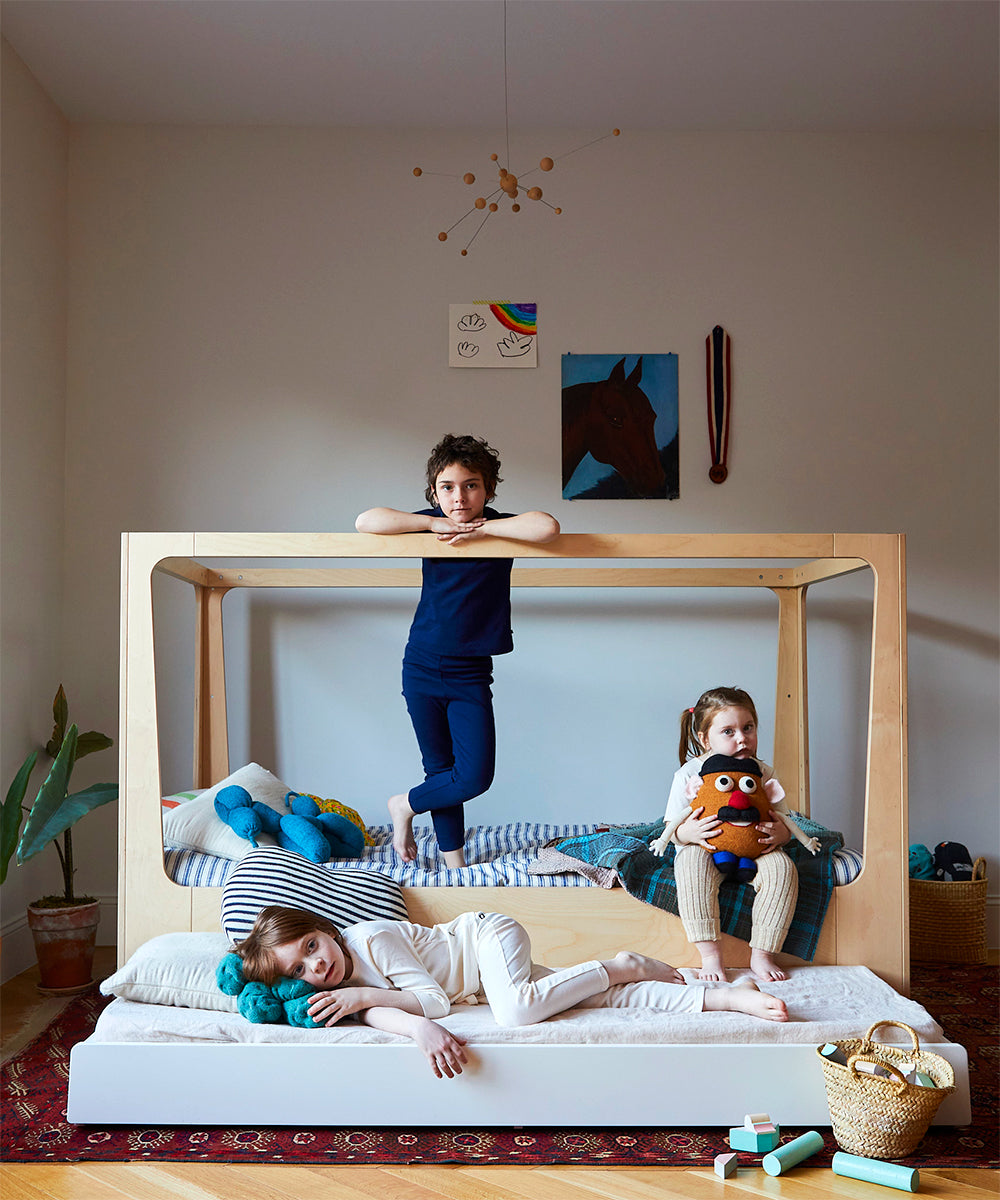 Three children pose in a stylish room featuring the Perch Nest Bed, surrounded by toys and colorful wall decor.