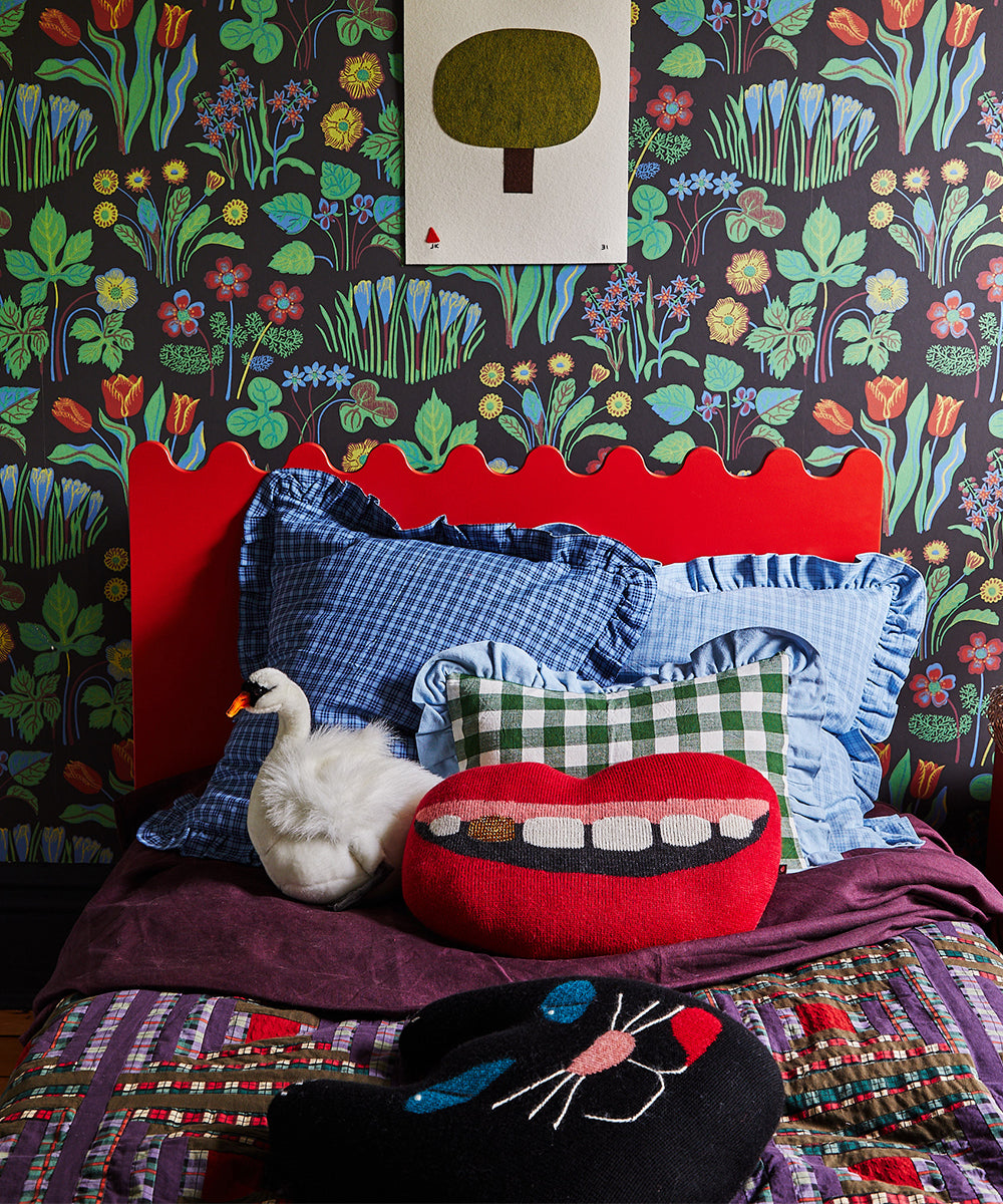 A colorful bed with a Black Cat Pillow, quirky cushions, floral wallpaper, and tree art above the headboard.