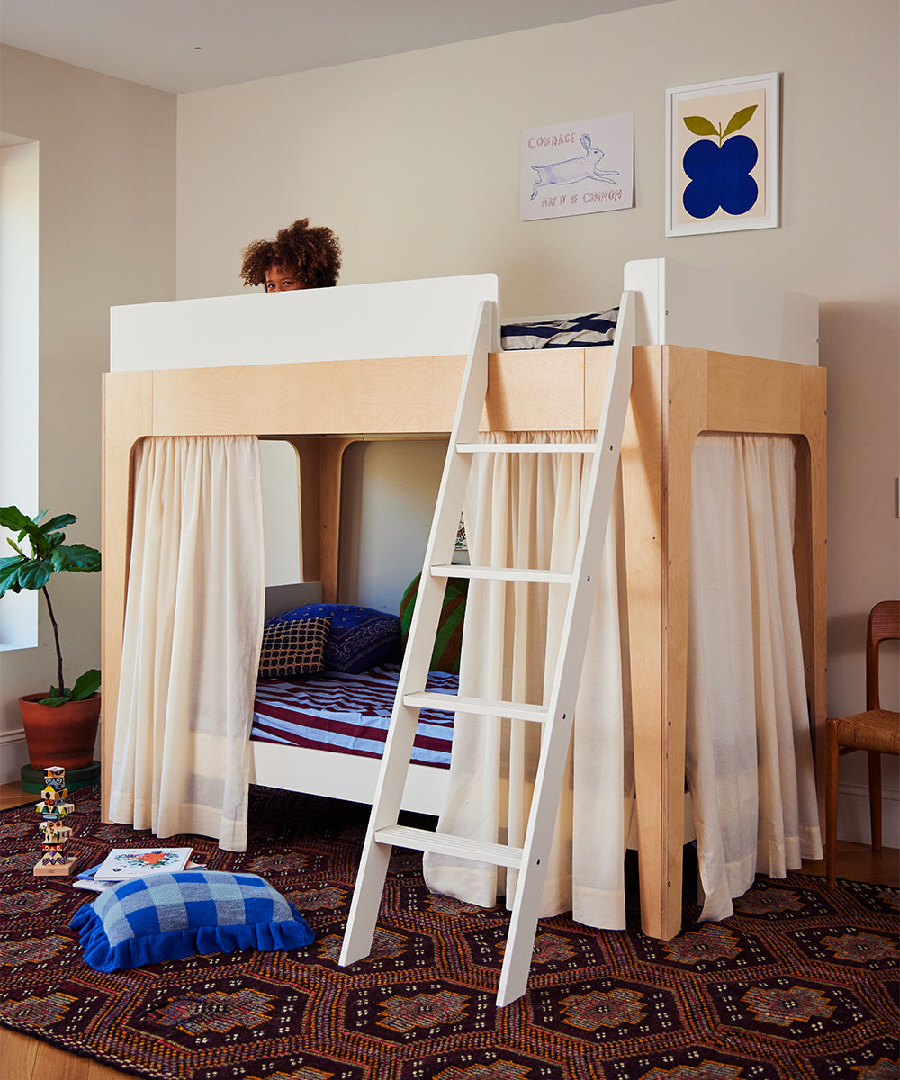 A childs head peeks over the top of the Perch Twin Bunk Bed, which has a ladder and curtains below, with pillows on a patterned rug. Wall art decorates the background.