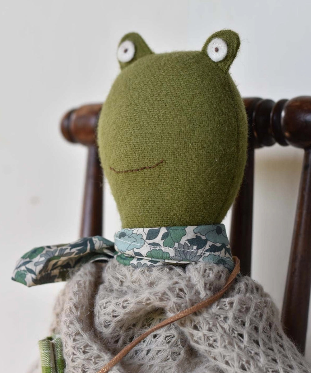 Fern le Frog, a plush frog doll with a floral scarf and knitted shawl, sits on a wooden chair.