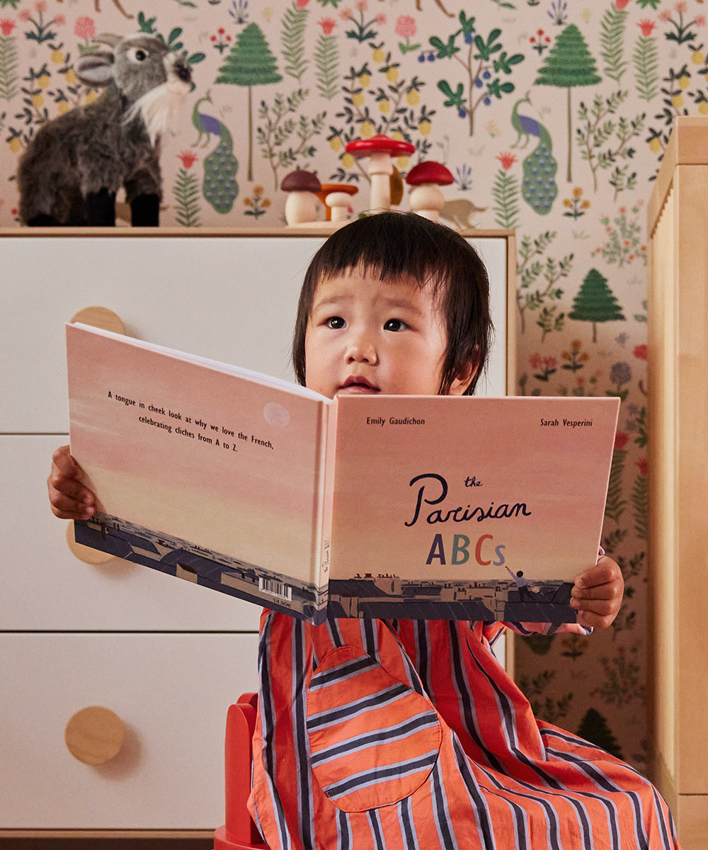 A child sits in a room with patterned wallpaper, holding and looking at an open book titled Le Parisian ABCs.