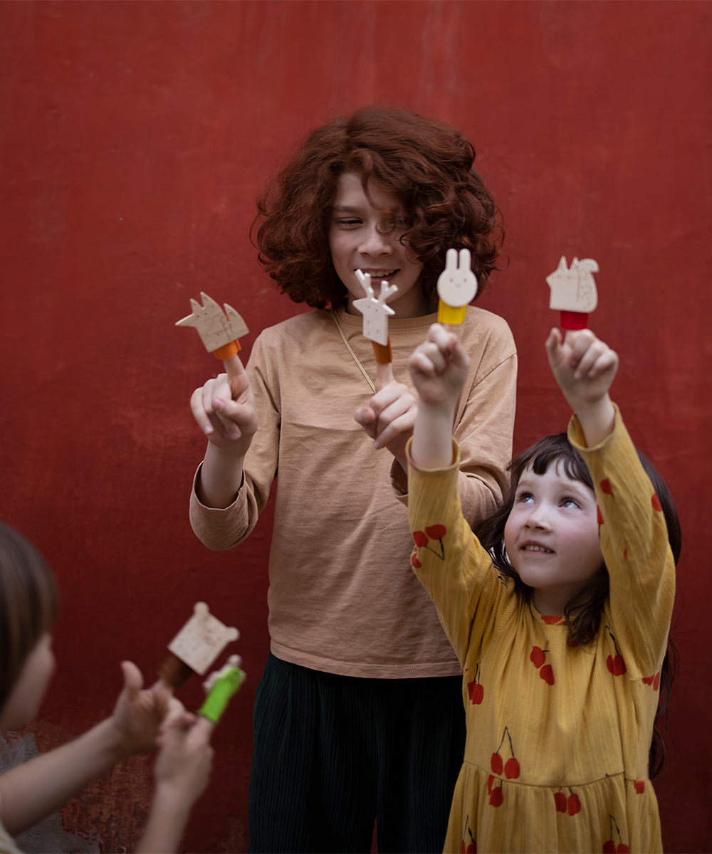 Two children joyfully hold animal-shaped ice creams against a red background; one is adorned in a beige shirt while the other twirls in a yellow dress, playing with the delightful Le Deer Finger Puppet.