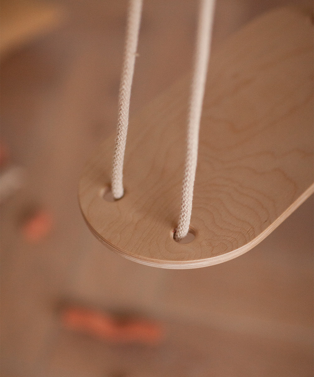 The image shows a Le Wooden Swing, featuring a wooden seat with two rope handles. The background is blurred.