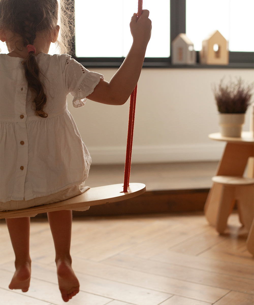 A young girl in a white dress sits on the Le Wooden Swing indoors, facing a window with wooden decor and a potted plant visible in the background.