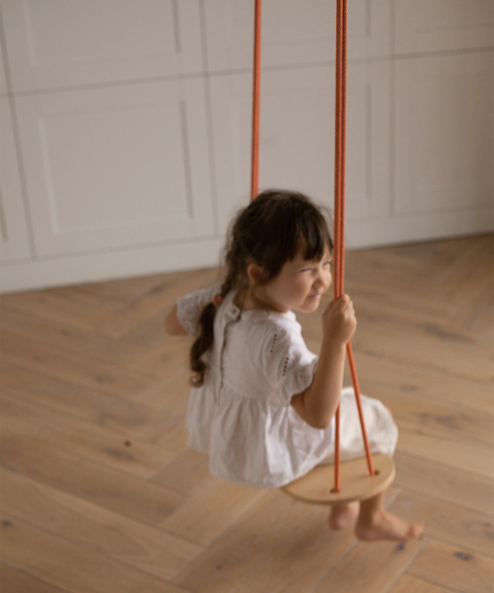 A child with braided hair sits on a Le Wooden Swing with orange ropes, indoors on a wooden floor, wearing a white dress.