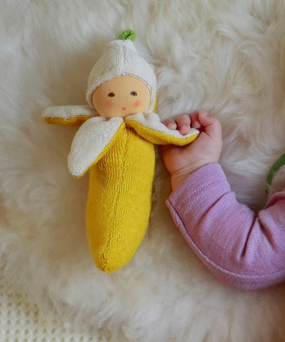 A baby’s hand holds the Banana Rattle, a soft banana-shaped toy with a smiling face, resting on a fluffy white surface.