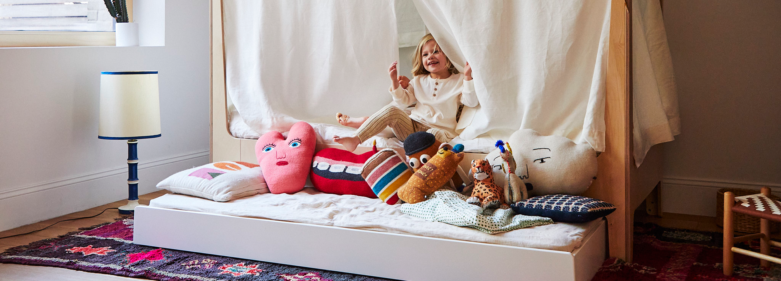 Child sitting in a cozy bed with white curtains, surrounded by colorful pillows and plush toys, smiling and holding a small object. A lamp and a patterned rug are visible in the room.