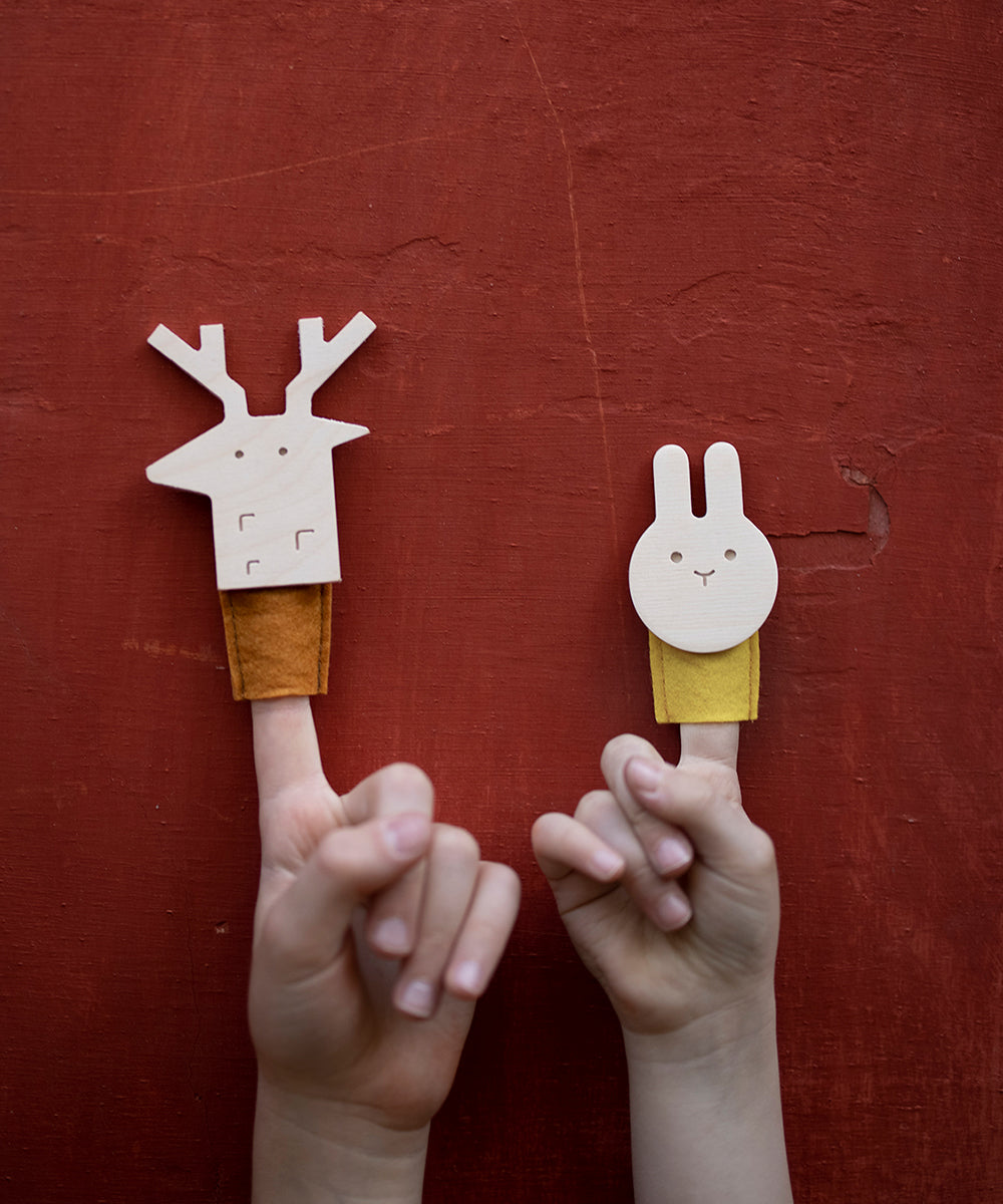 Two hands with finger puppets against a red background: Le Deer Finger Puppet on the left and a wooden rabbit on the right.