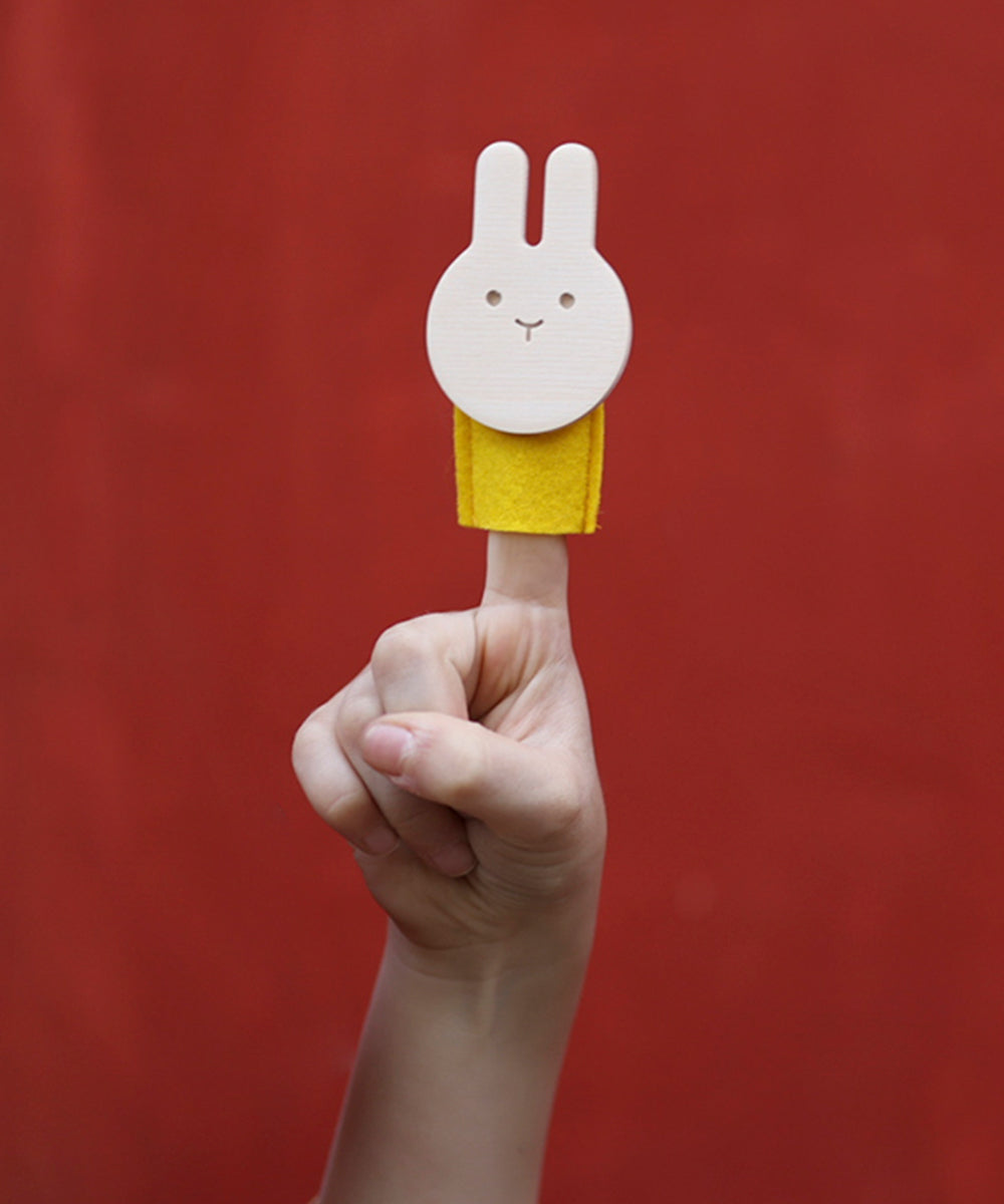 The Le Rabbit Finger Puppet, featuring a white rabbit with a yellow body, is displayed on a persons finger against a red background.