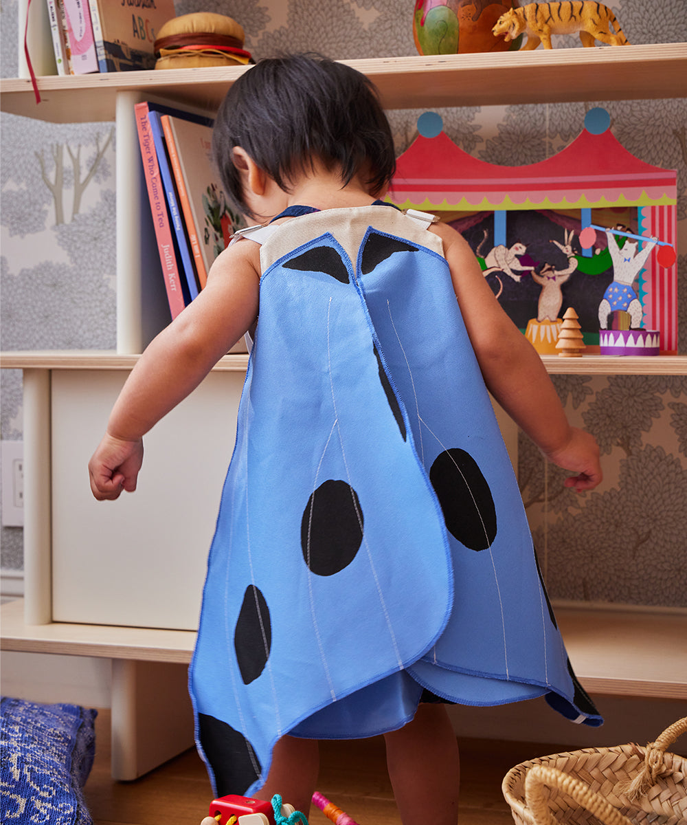 A toddler in the Le Blue Butterfly Wings Costume stands with arms outstretched in a colorful playroom.