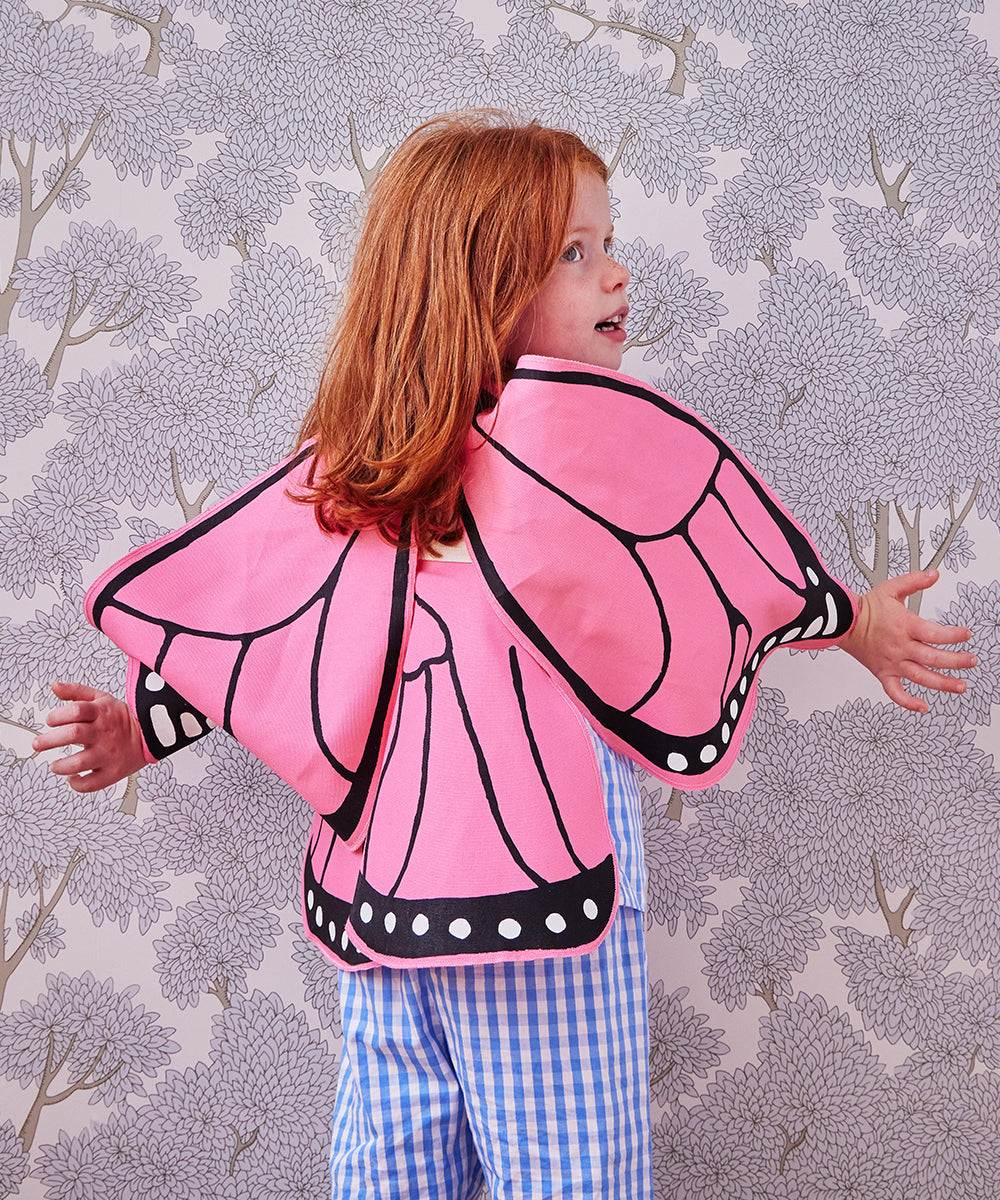 A child with red hair is wearing Le Pink Monarch Butterfly Wings and blue checkered pants, standing against floral-patterned wallpaper.