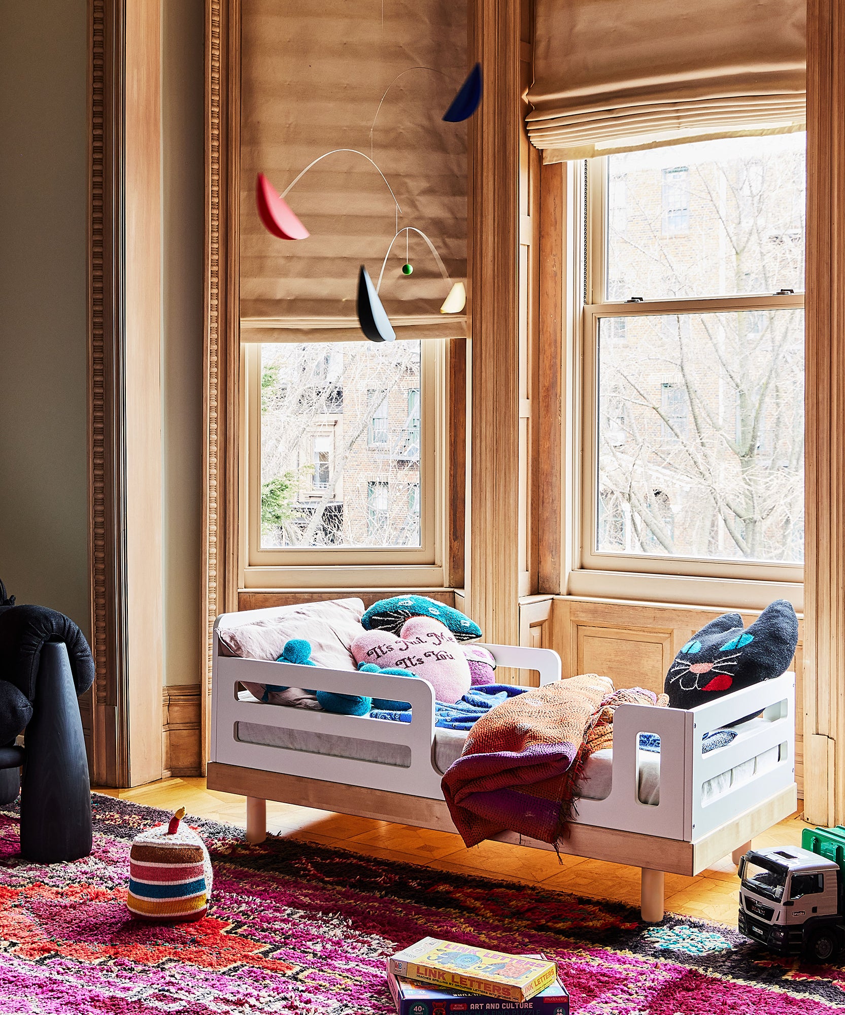 The Classic Toddler Bed, adorned with colorful pillows and blankets, is placed near large windows beside a bright, patterned rug.