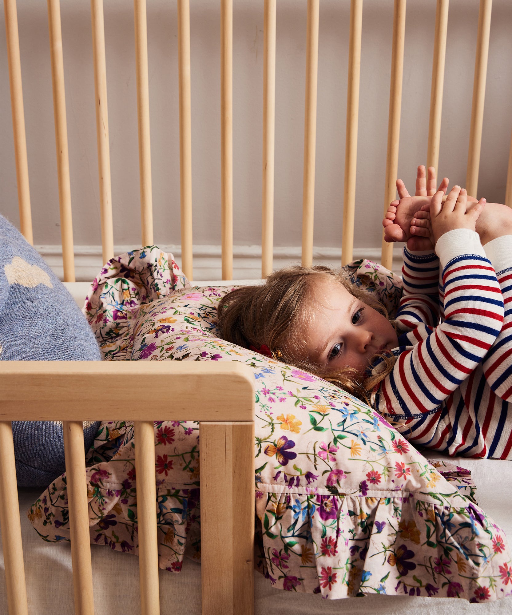A child in striped pajamas lies in a crib made with the Moss Crib Conversion Kit, holding their feet on a floral pillow.